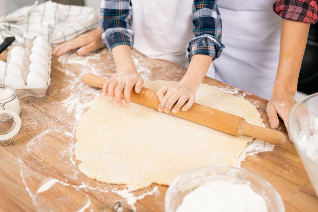 hands of child with wooden pin rolling fresh dough on table while helping mom